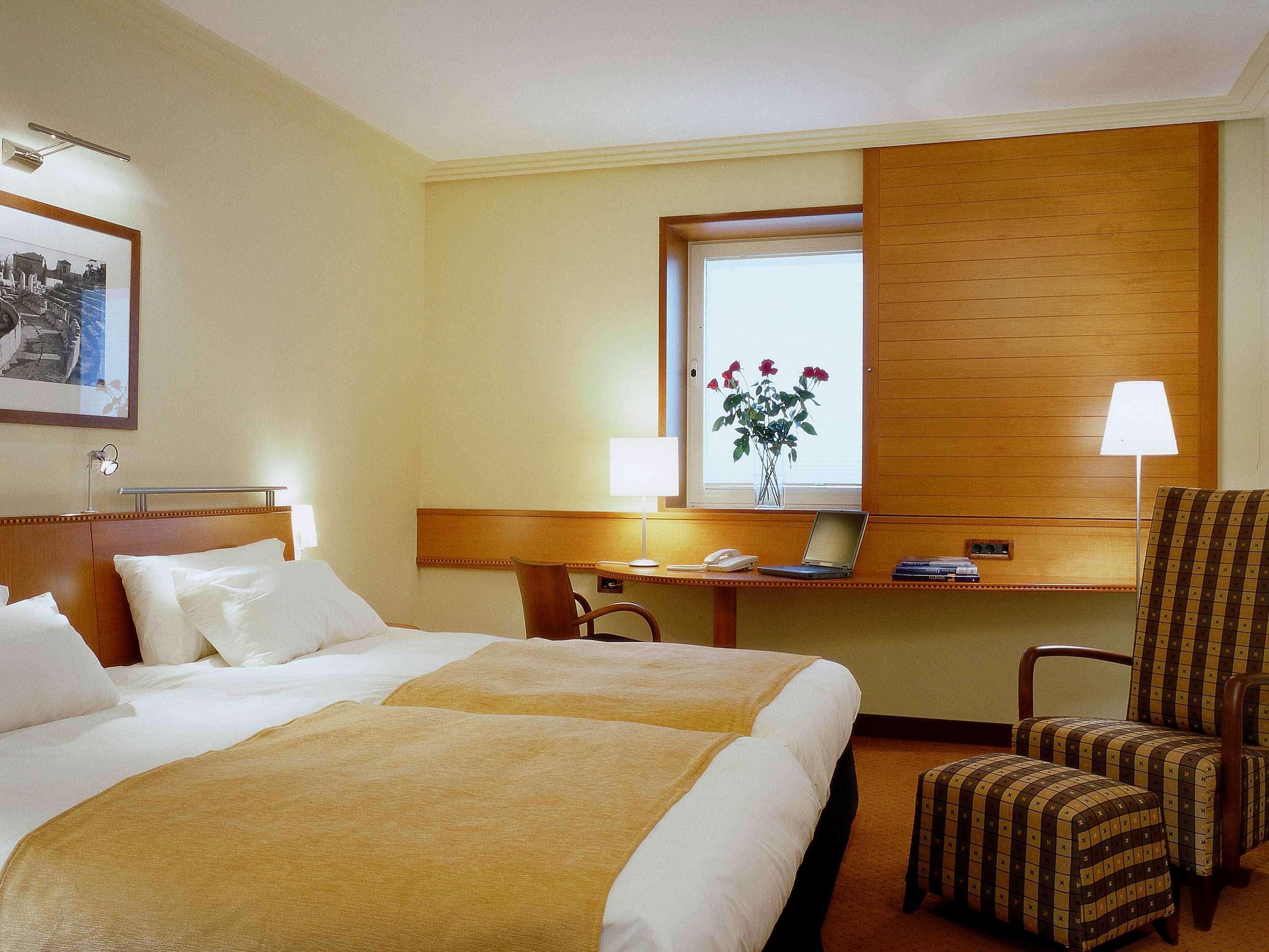 雅典索菲特机场酒店 Spáta 外观 照片 The photo shows a cozy hotel room. It features two twin beds with light-colored bedding and taupe blankets. There is a wooden headboard above the beds. A window with light drapes allows natural light to enter, and there are decorative roses on the wi