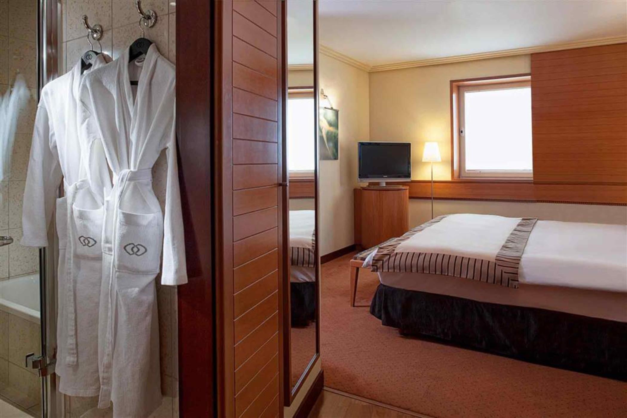 雅典索菲特机场酒店 Spáta 外观 照片 The photo depicts a cozy hotel room. On one side, there's a set of wooden wardrobes, and hanging from one of them is a white bathrobe. The room features a neatly made bed with a striped blanket and decorative pillows. There's a small television mount