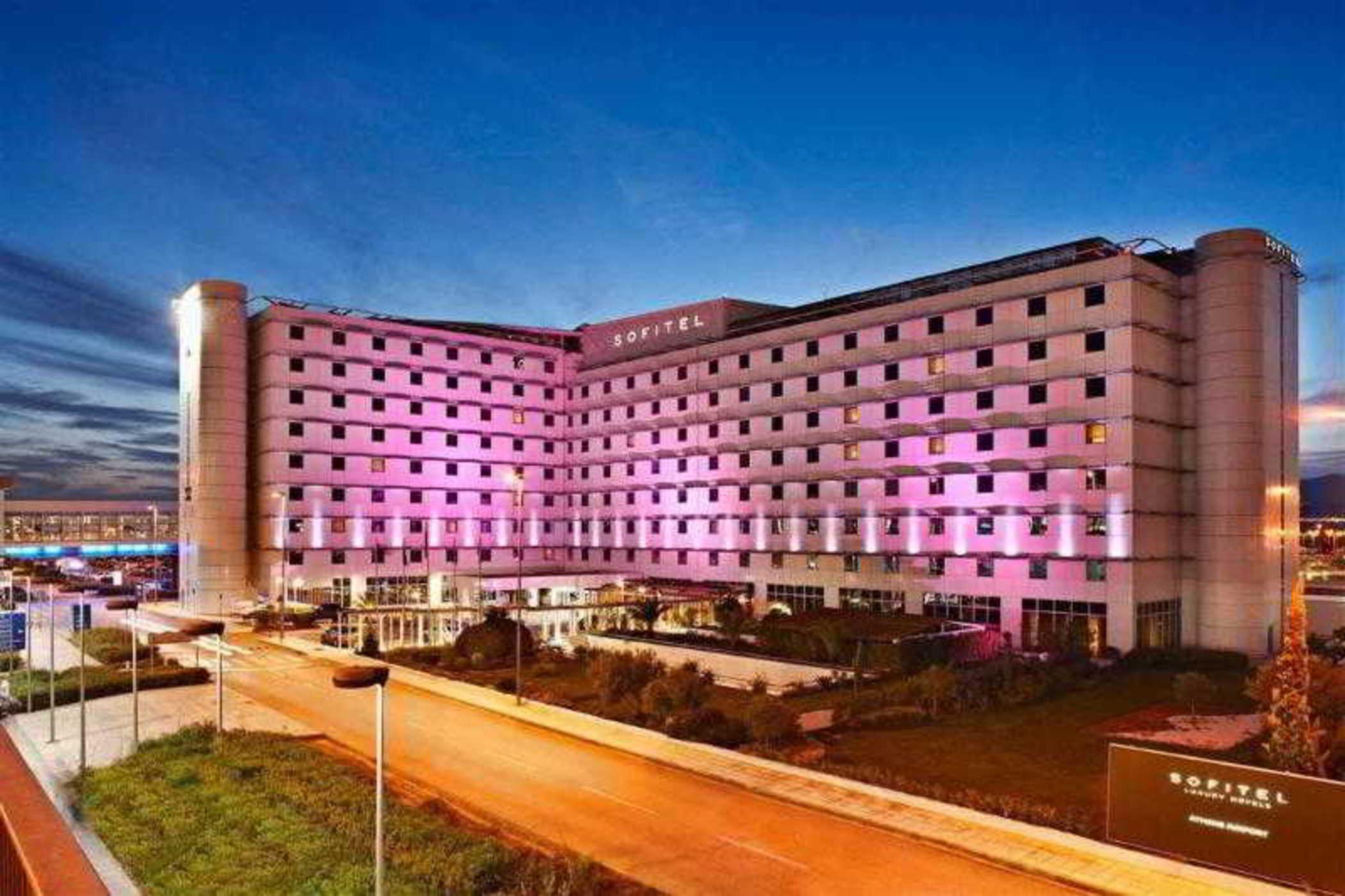 雅典索菲特机场酒店 Spáta 外观 照片 The photo displays a modern hotel building, likely a hotel brand, with a sleek, contemporary architectural design. The façade is illuminated in pink and purple lights, suggesting a vibrant atmosphere. The building has multiple floors, with large wind