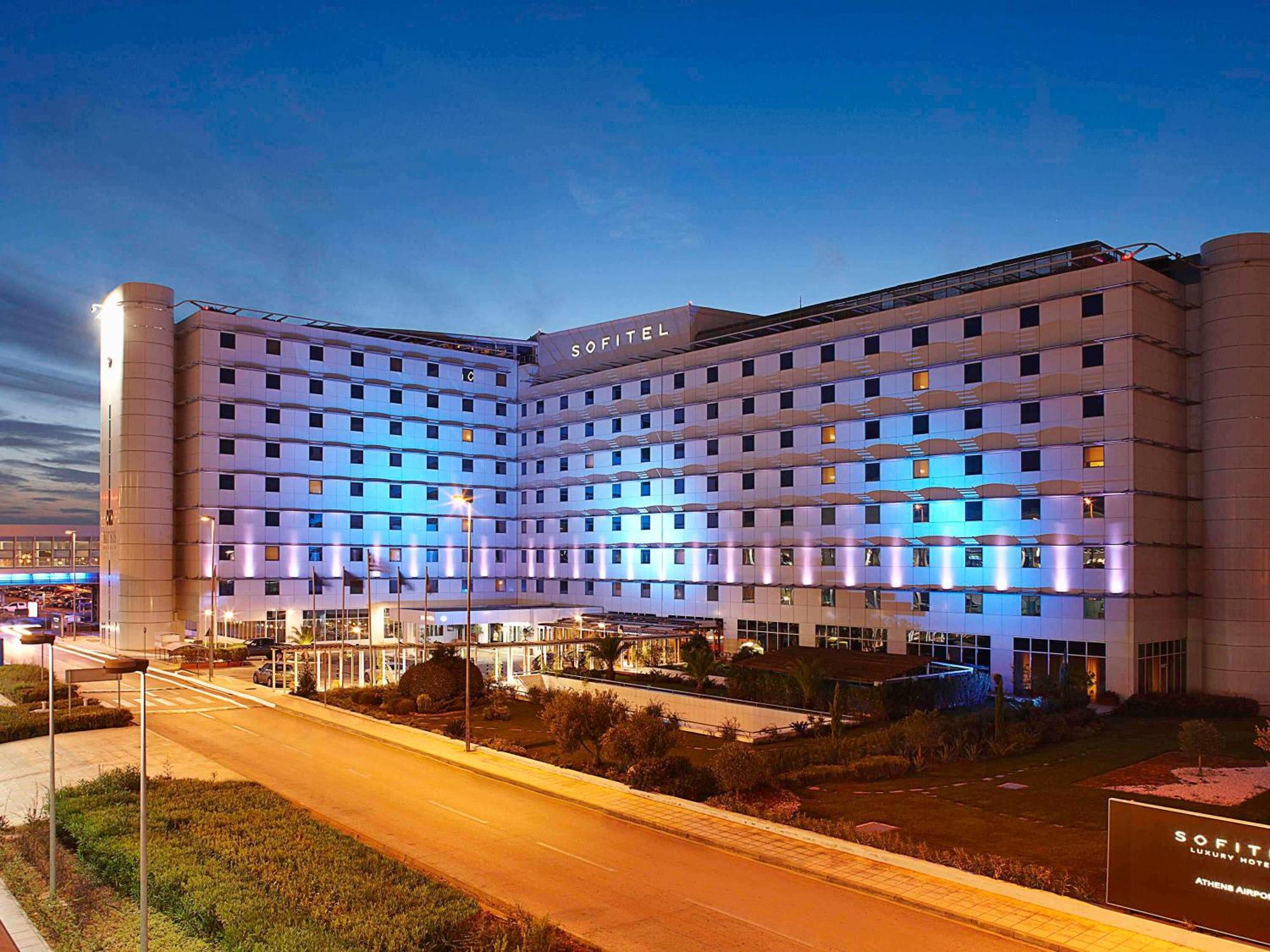 雅典索菲特机场酒店 Spáta 外观 照片 The photo shows a large modern hotel building, likely named "Sofitel," illuminated with blue and purple lights as evening approaches. The structure features multiple floors and a contemporary design, with large windows and a flat facade. In front of 